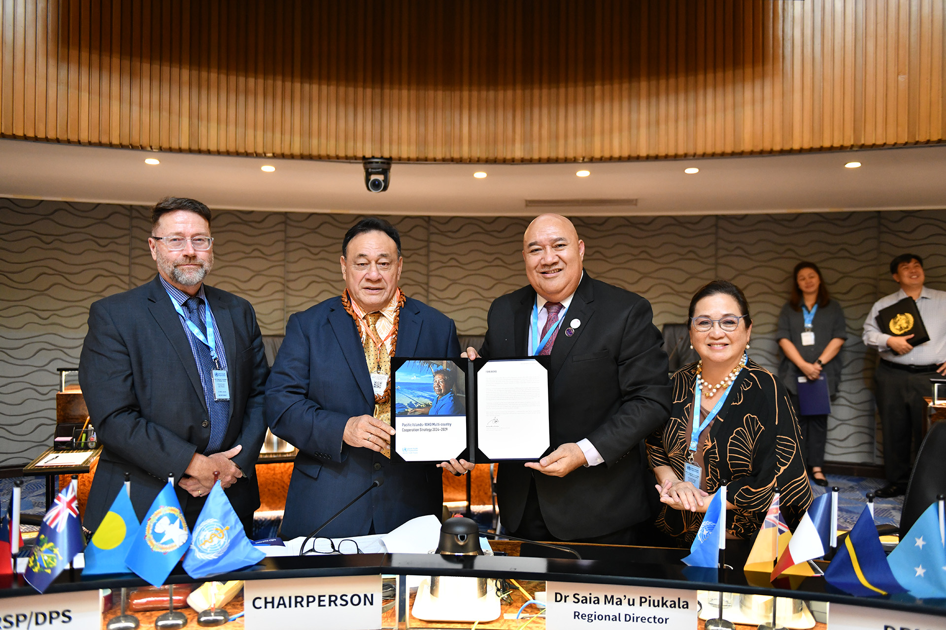 Four individuals stand behind a table during an international event in a conference room, dressed formally. Two hold a document titled 'Pacific Islands: WHO Multi-Country Cooperation Strategy 2024-2029
